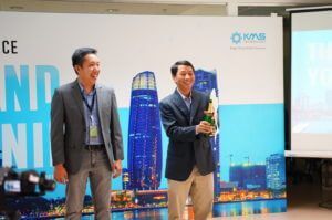 Two men stand in front of a banner with the KMS Logo and Da Nang Skyline. Both men are smiling and one is holding a bottle of champaign. 
