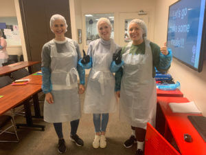 Three women stand smiling, wearing white plastic aprons, blue plastic arm covers, and hairnets. The two on the right are giving a thumbs up.