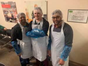 Three men stand smiling, wearing white plastic aprons, blue plastic arm covers, and hairnets. The man in the middle has his arms crossed.
