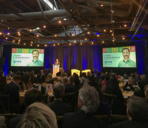 This image shows the award ceremony for the Leaders in Corporate Citizenship Award. A man stands on stage behind a podium addressing the crowd. To his left and right are two screens with Josh's photo and the KMS logo. 